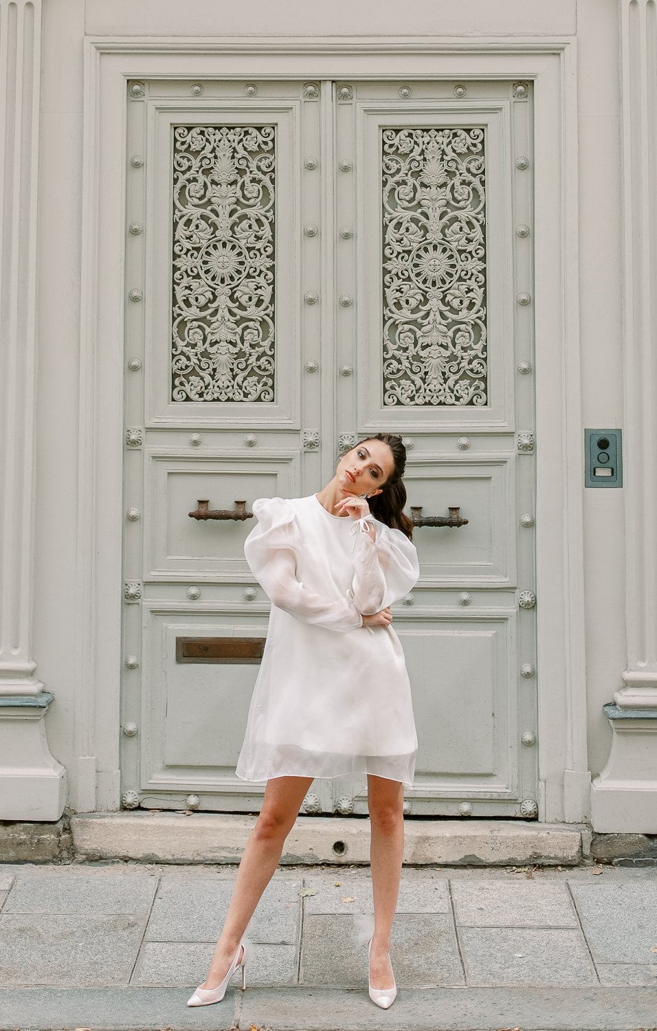 Bride in short white puff-sleeve dress posing before an ornate Parisian doorway in soft morning light