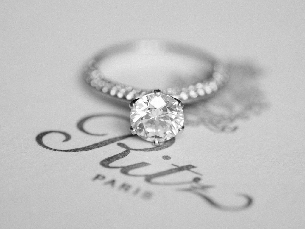 Diamond solitaire engagement ring on pave band resting on Ritz Paris stationery in black and white