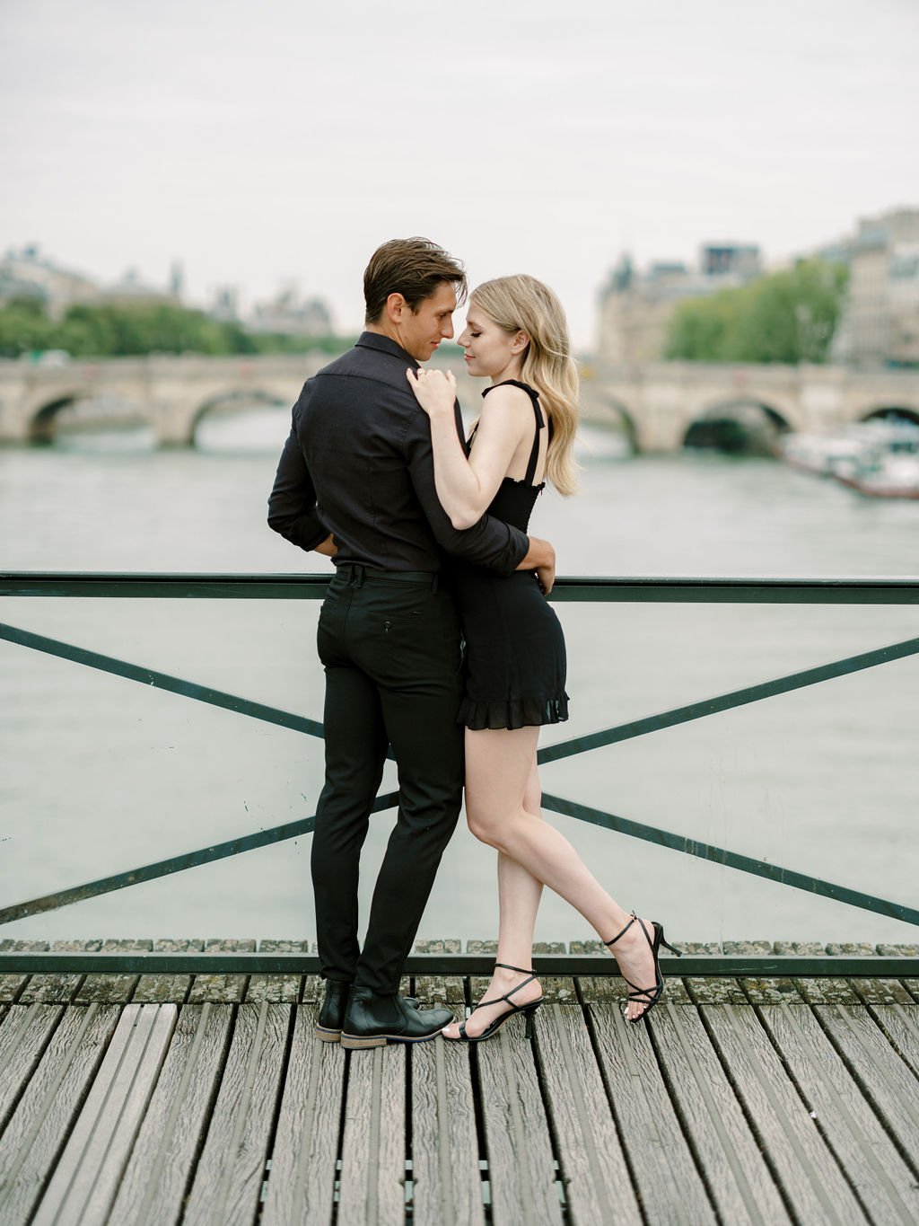 Couple in black embracing on Pont des Arts bridge with Seine River and Parisian stone bridge in soft overcast light