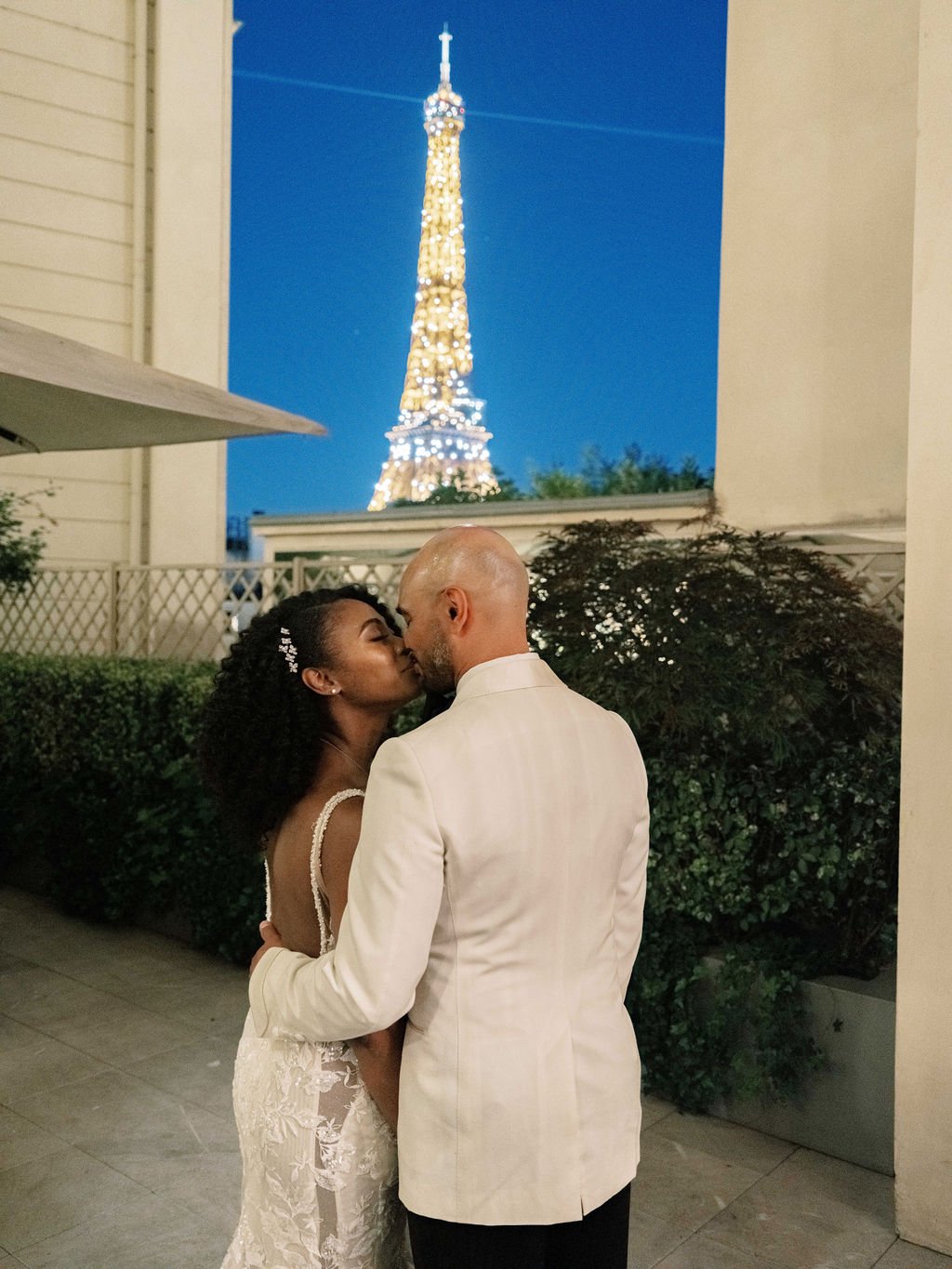Couple kissing at Shangri-La Paris terrace with sparkling Eiffel Tower illuminated against deep blue twilight sky