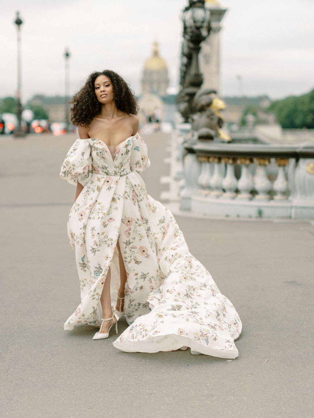 Bride in flowing floral off-shoulder gown on Pont Alexandre III with gilded statues and Les Invalides dome behind her