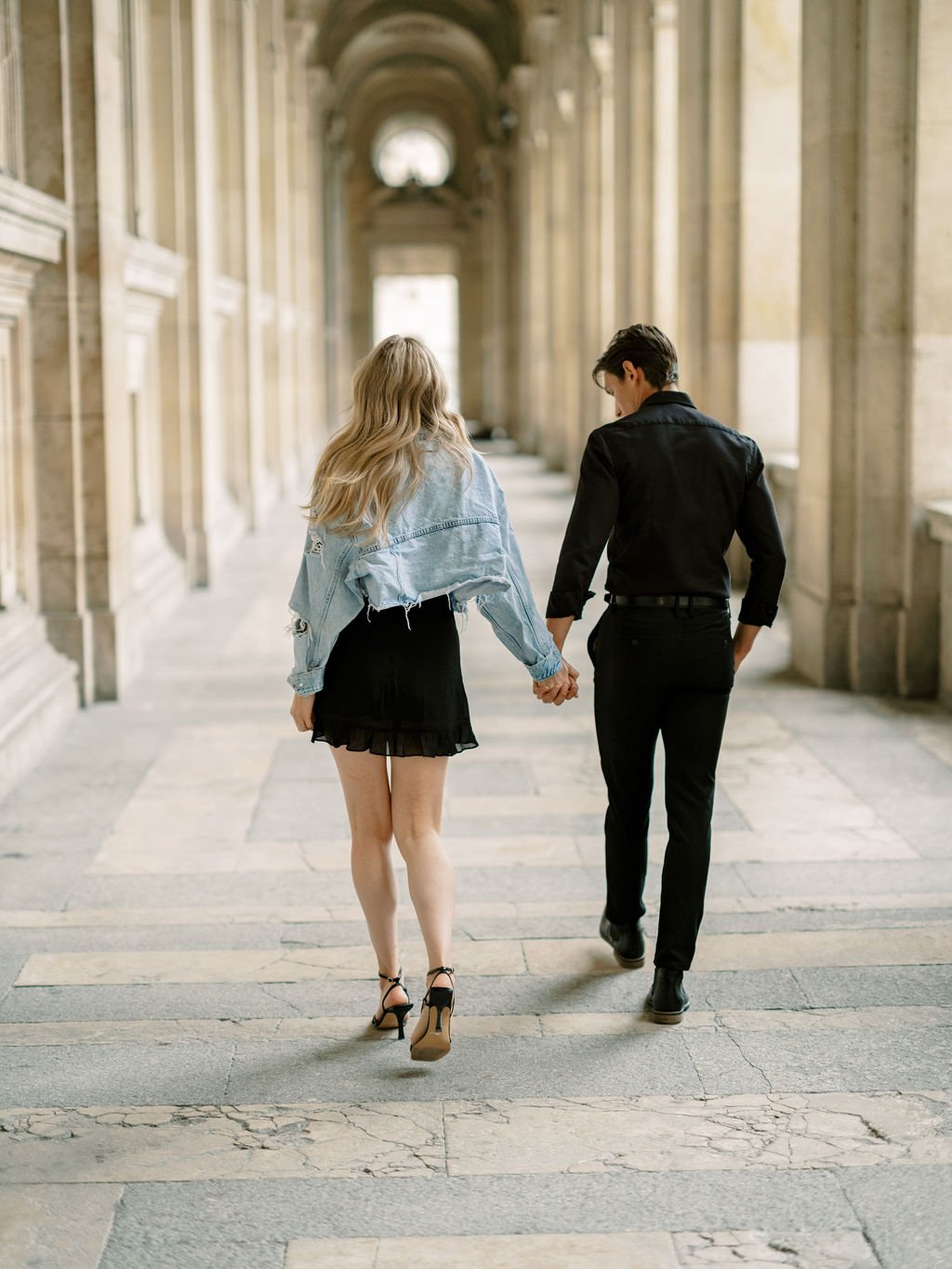 Couple in casual chic outfits walking through the Louvre colonnade during a Paris engagement session