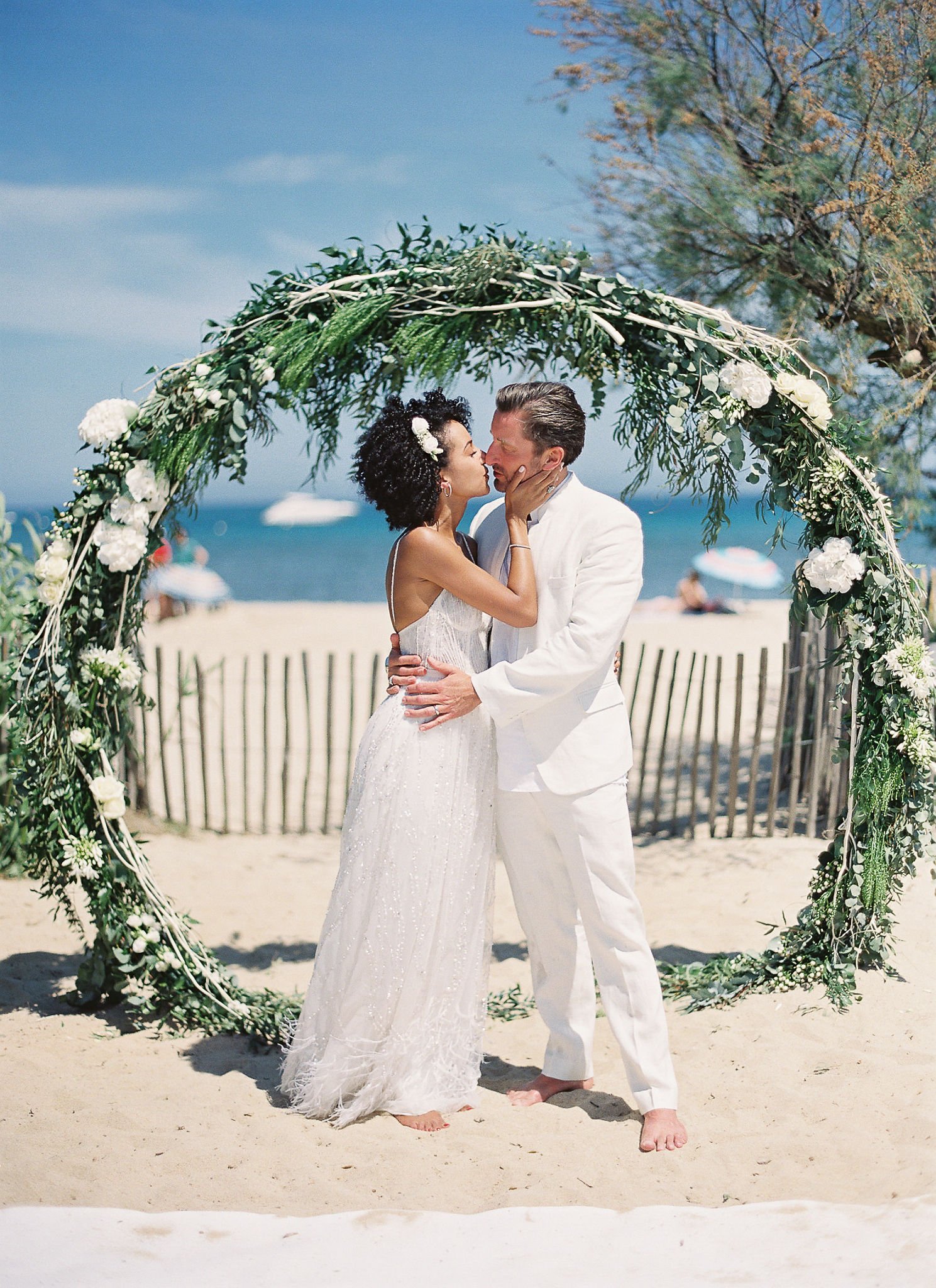 Couple sharing their first kiss under a eucalyptus arch on Saint-Tropez beach in golden afternoon light
