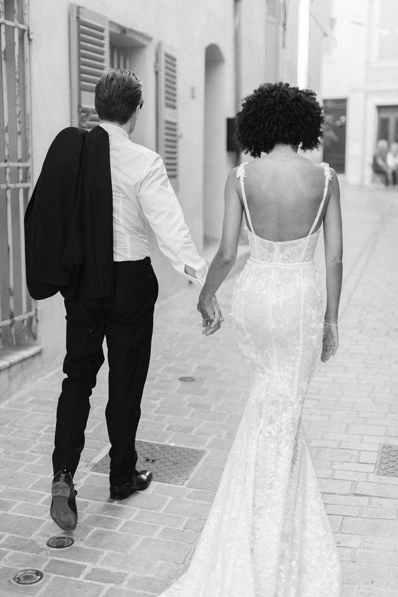 Newlywed couple walking hand-in-hand down cobblestone streets in Saint-Tropez, bride in backless gown