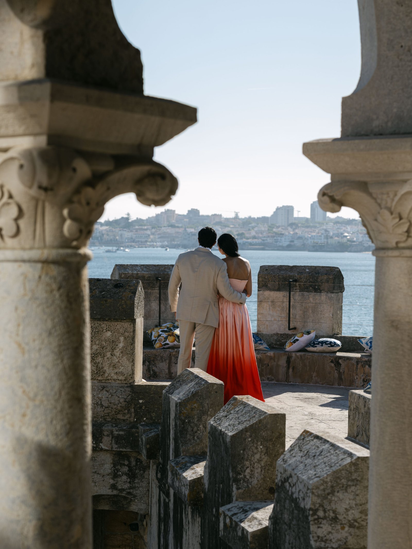 Indian wedding couple embracing between the stone columns of Belém Tower, Lisbon, with the Tagus river behind