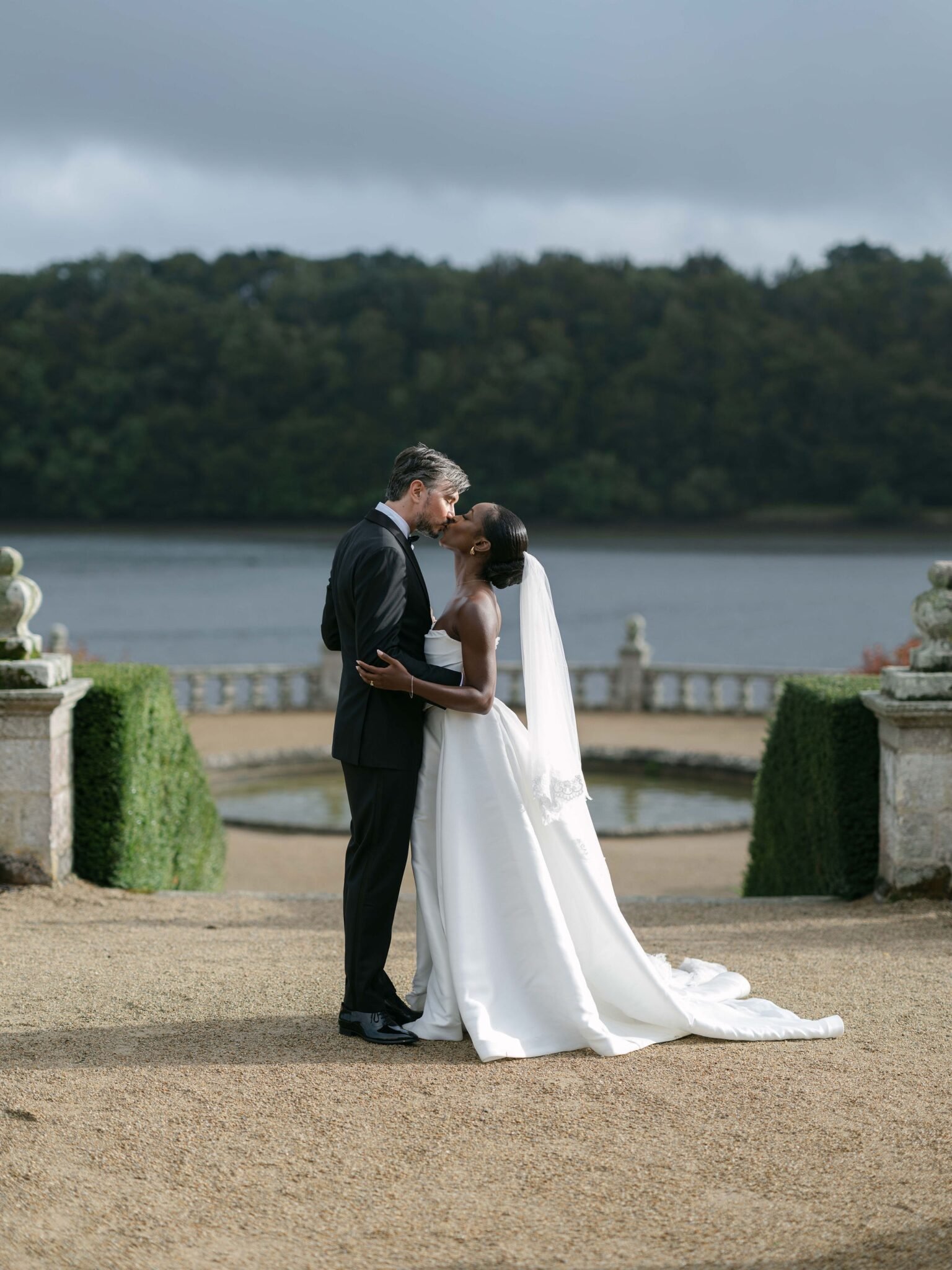 Bride and groom kissing on a stone terrace over the water during an Orangerie de Lanniron wedding under a dramatic Brittany sky