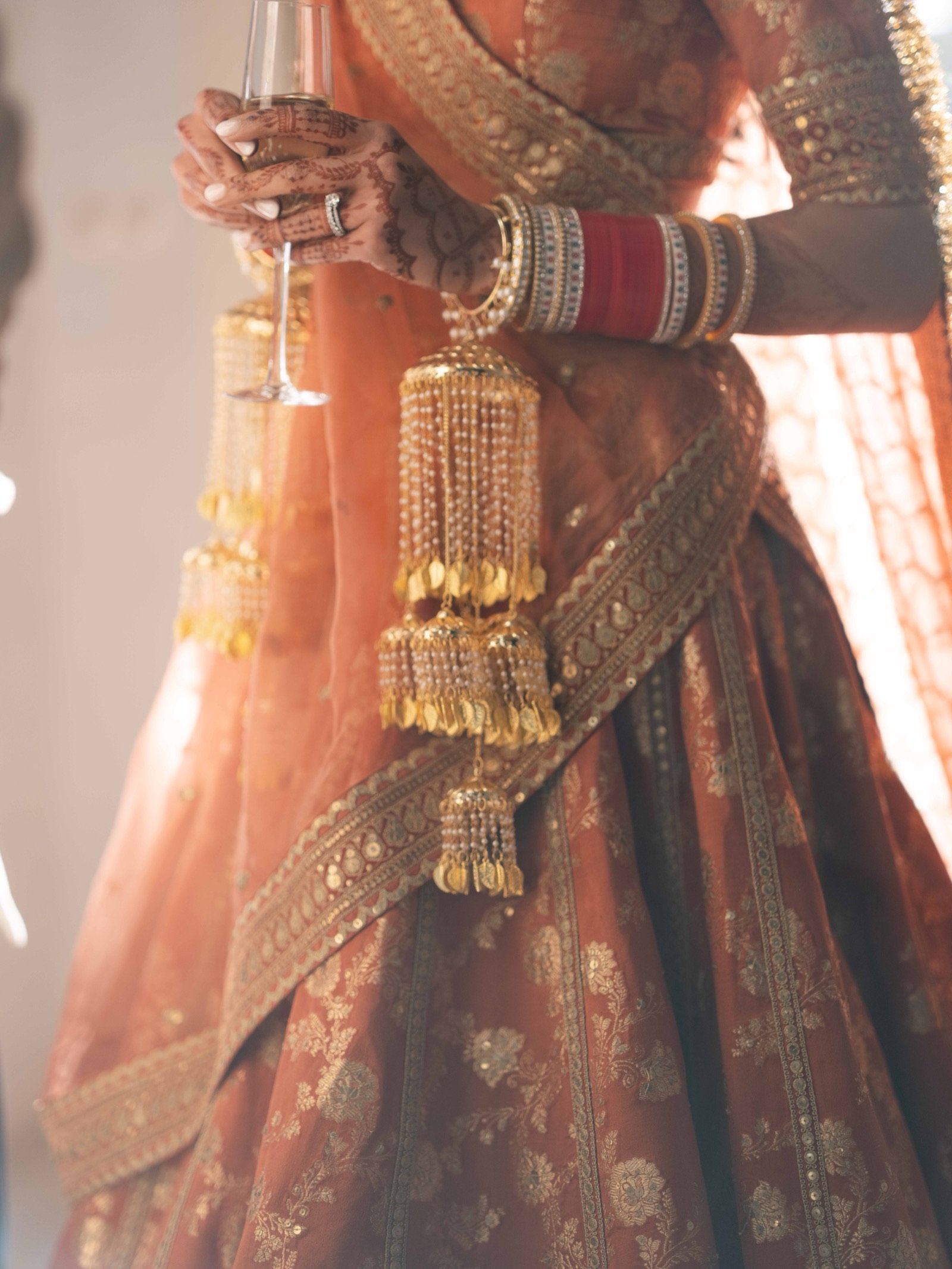 Bride in a coral and gold lehenga holding a glass of champagne, mehndi on her hands and kaleere at her wrist, in warm evening light in Lisbon
